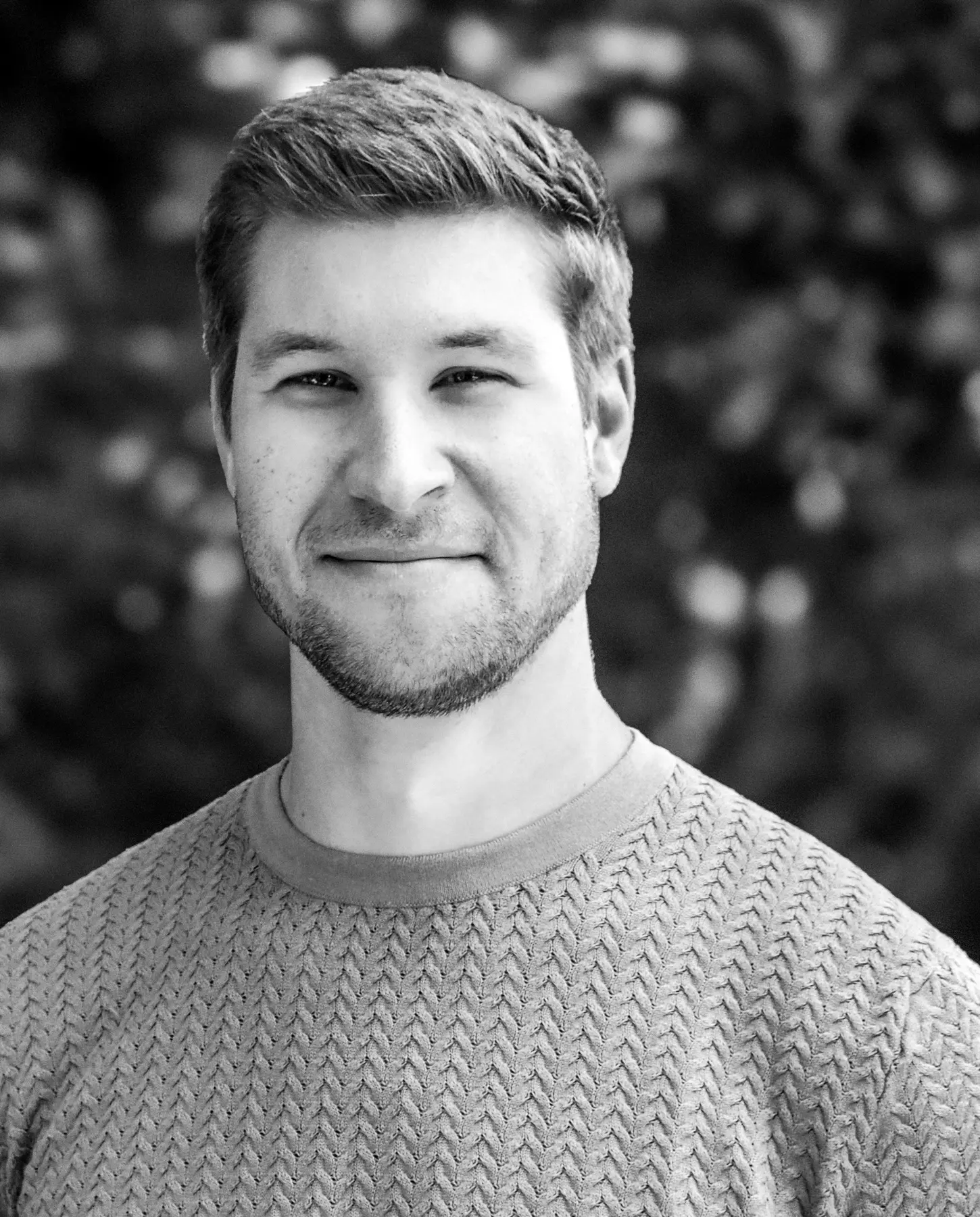 Black and white headshot of a young man with short hair and a beard, smiling, wearing a textured crewneck sweater
